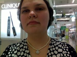 A picture of me wearing a delicate single strand of pearls that has a single silver flower in the middle. The flower is about twice the width of the necklace with inward-curling petals. In the centre of the flower is a single stone that catches the light. 