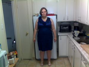 A picture of me as I'm about to depart for a wedding. I am wearing a sleeveless, cobalt blue cocktail dress that ends just above my knees. The dress has a straight cut and has vertical seams down the front. It is made of a shiny fabric that gives what someone called an "icy cast" to the blue shade. There is a ruffle detail that moves diagonally over my right shoulder and terminates in a stylized flower on the right side of the v-neckline. My hair has been styled into a moere voluminous bob. I have matched the dress with sparkly silver sandals, a silver satin clutch, a tripple strand of pearls with the odd silver bit in them and diamond stud earrings.   
